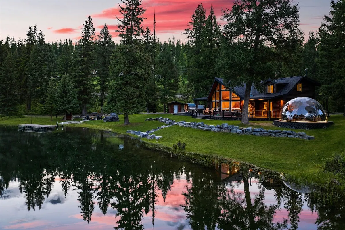 The cabin at Lodge 93 across the river channel at sunset, its windows and geodome glowing, the whole sky mirrored in the water